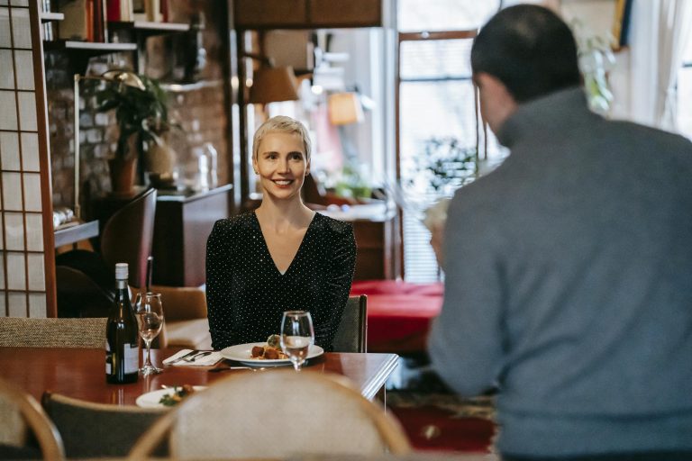 Positive adult couple having date in bright room while man standing with bouquet with flowers near chairs and table with glasses and wine bottle near plate with food and cutlery