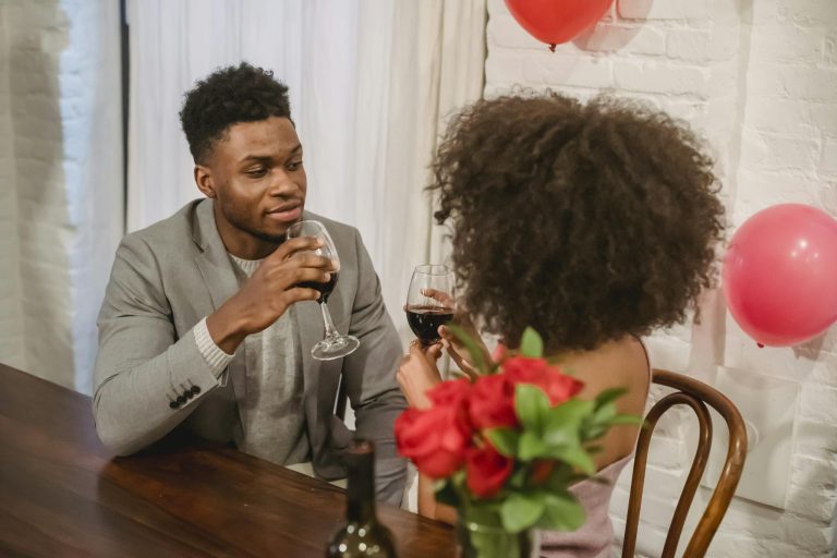 Young black couple in elegant clothes sitting at table while drinking wine from glasses near bottle and vase with flowers bouquet in loft with balloons while looking at each other